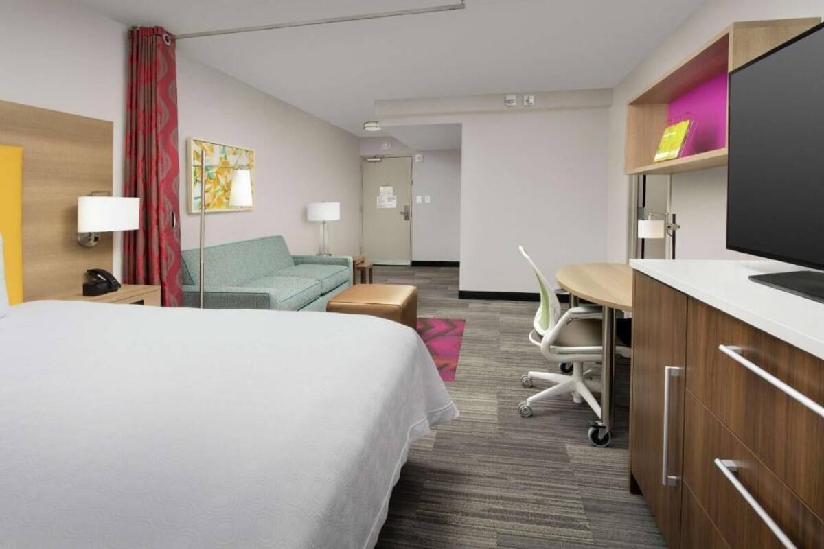 A hotel room with a bed, a desk, a chair, a sofa, a TV, and a desk lamp.