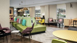 A modern lounge area with a green sofa, yellow and purple chairs, a round table, and large windows providing a view of the outside. - Home2 Suites by Hilton Atlanta Midtown (Atlanta)
