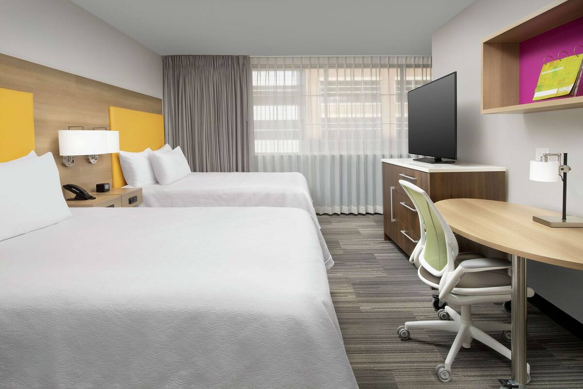 A hotel room with two beds, a desk, a TV, and a window with curtains.