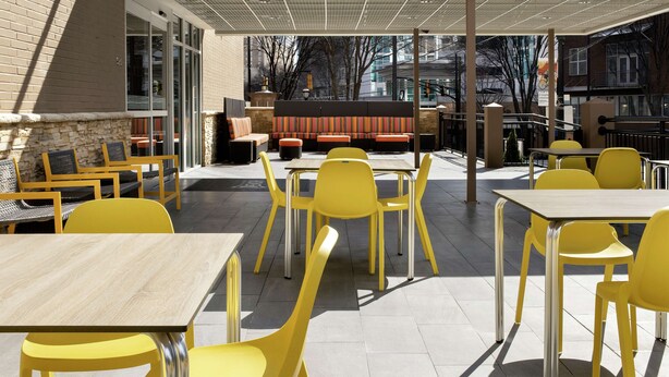 An outdoor patio with yellow chairs and tables, a brick wall, and a seating area with orange cushions. - Home2 Suites by Hilton Atlanta Midtown (Atlanta)