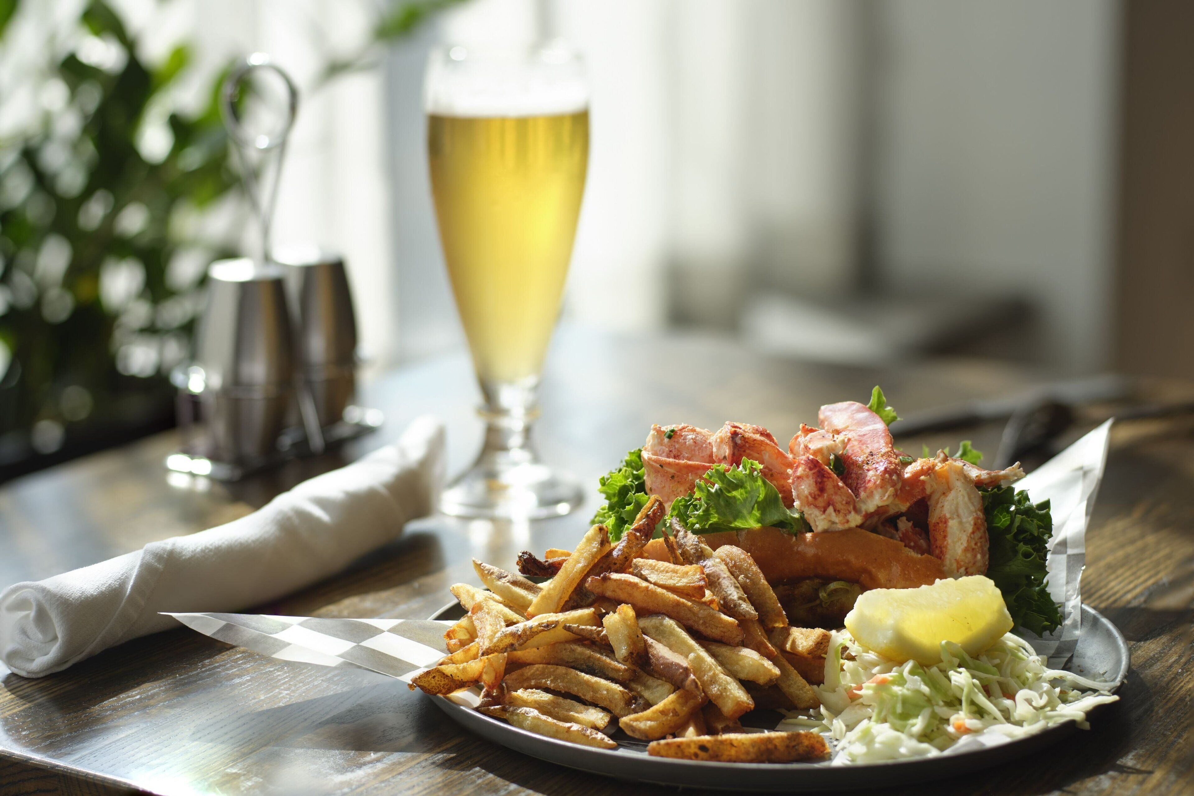 Ein Teller Essen mit Pommes, einem Lobster Roll und einem Bierglas auf einer Holztischplatte.