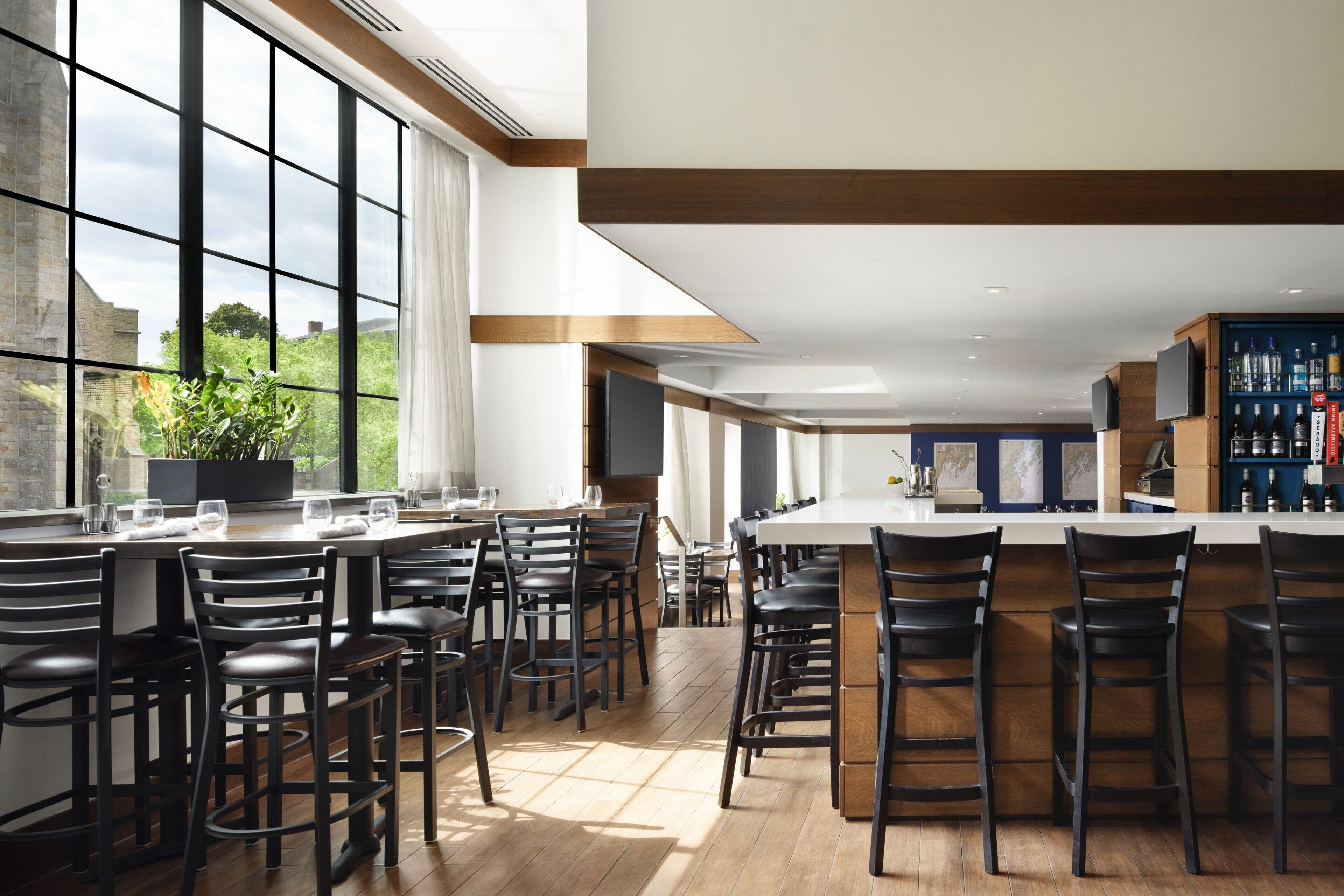 A modern bar area with high ceilings, large windows, and a view of greenery outside.