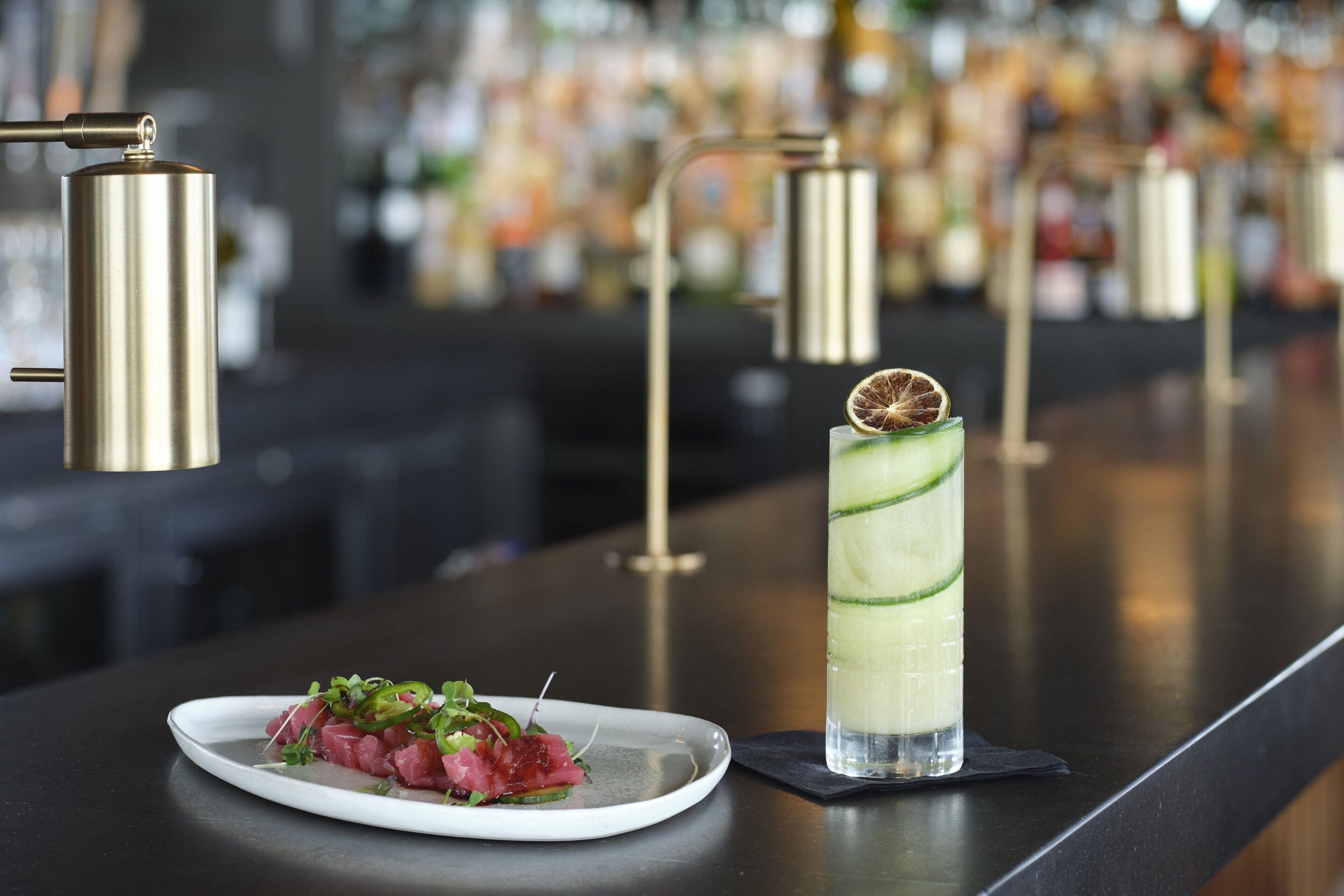 Une assiette blanche remplie de nourriture est posée sur un comptoir de bar, à côté d’un grand verre contenant une boisson verte.