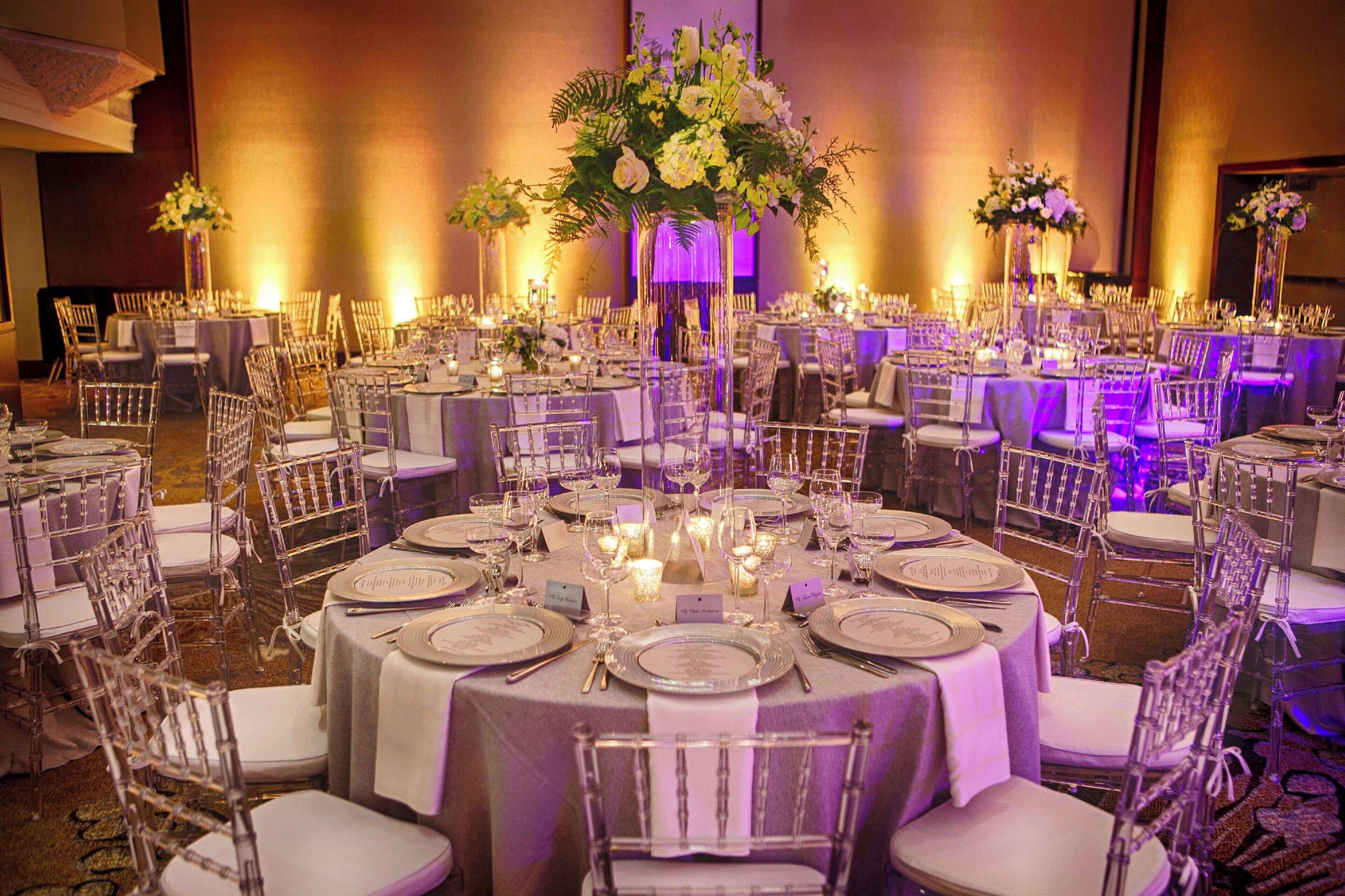 A banquet hall with round tables, clear chairs, and floral centerpieces.