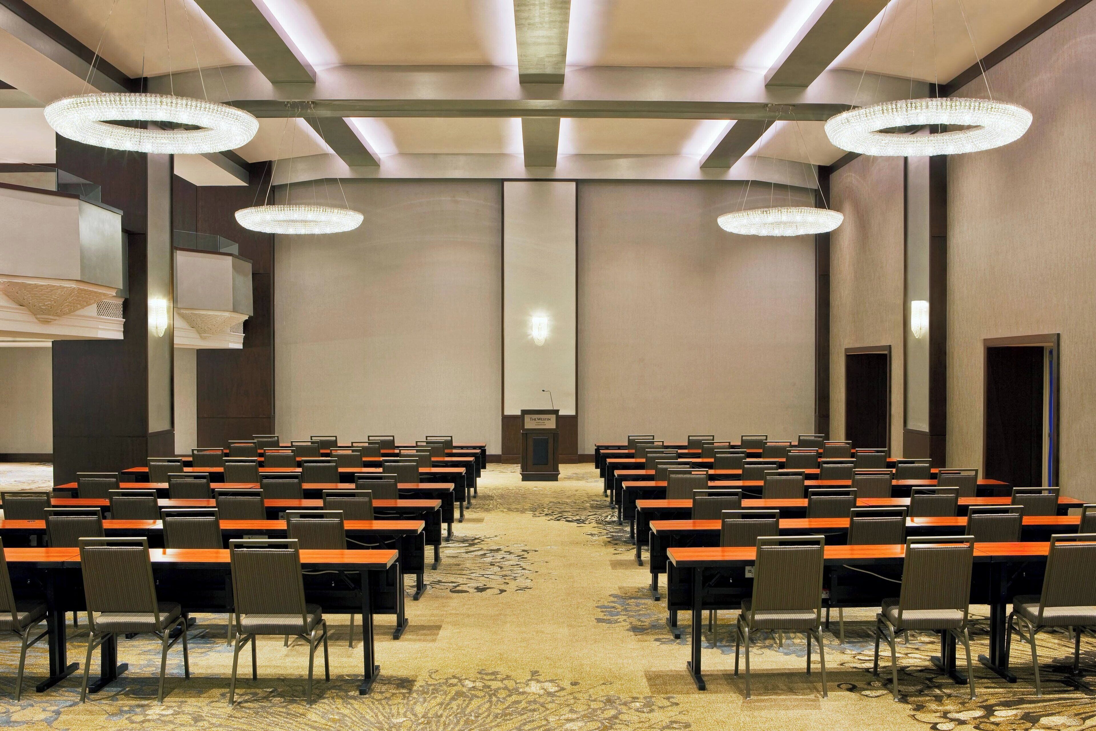 A conference room with rows of tables and chairs, a podium at the front, and chandeliers.