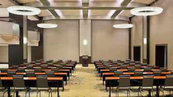 Uma sala de conferências com fileiras de mesas e cadeiras, um estrado à frente e lustres.