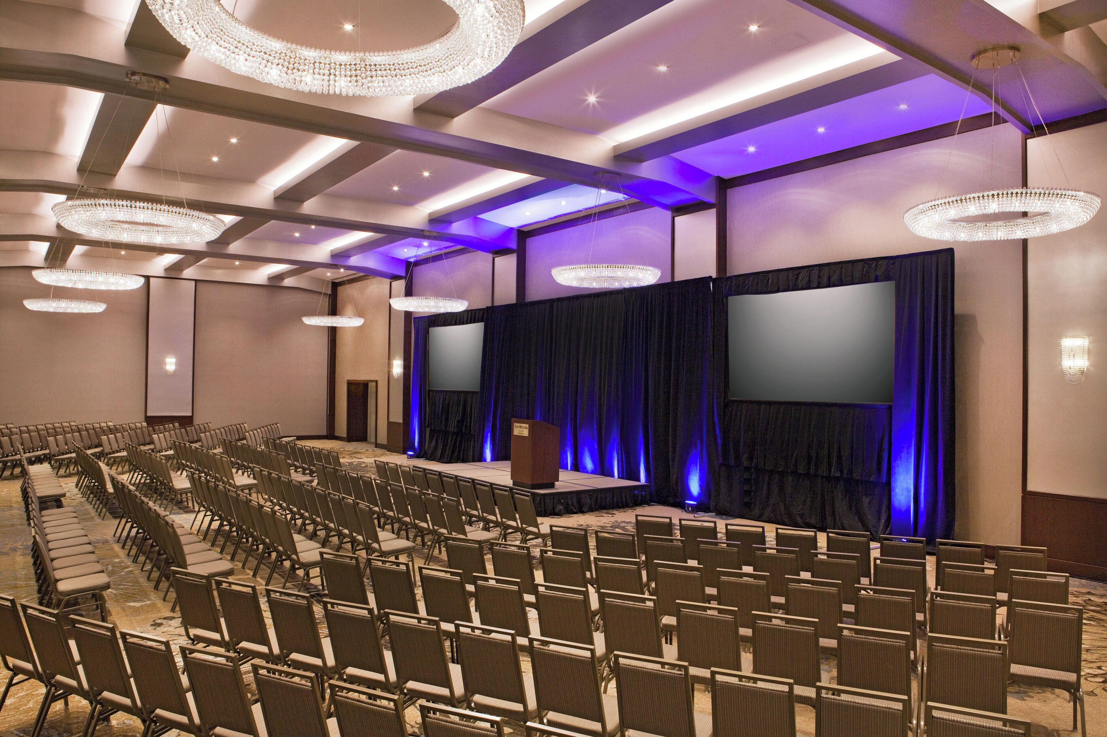 Uma sala de conferências com fileiras de cadeiras, palco com cortinas e lustres.