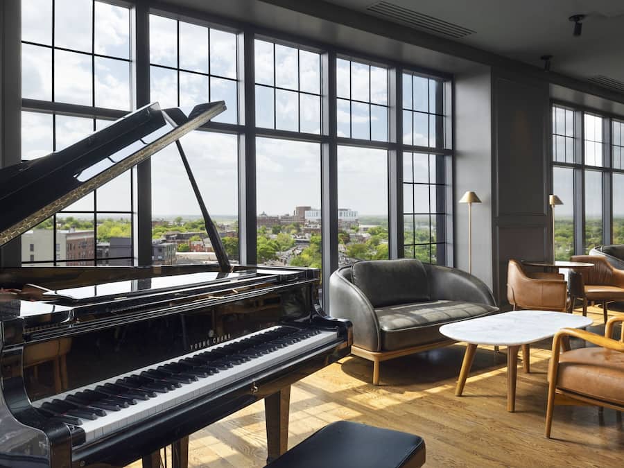 Un piano de cola en una habitación amplia con ventanas grandes, sillones de cuero y una pequeña mesa.