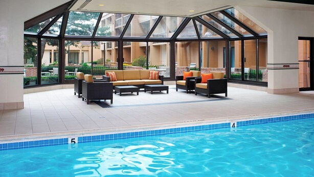 Un espace piscine doté d’un patio fermé par des baies vitrées, de coins salon extérieurs et offrant une vue dégagée sur les bâtiments environnants.