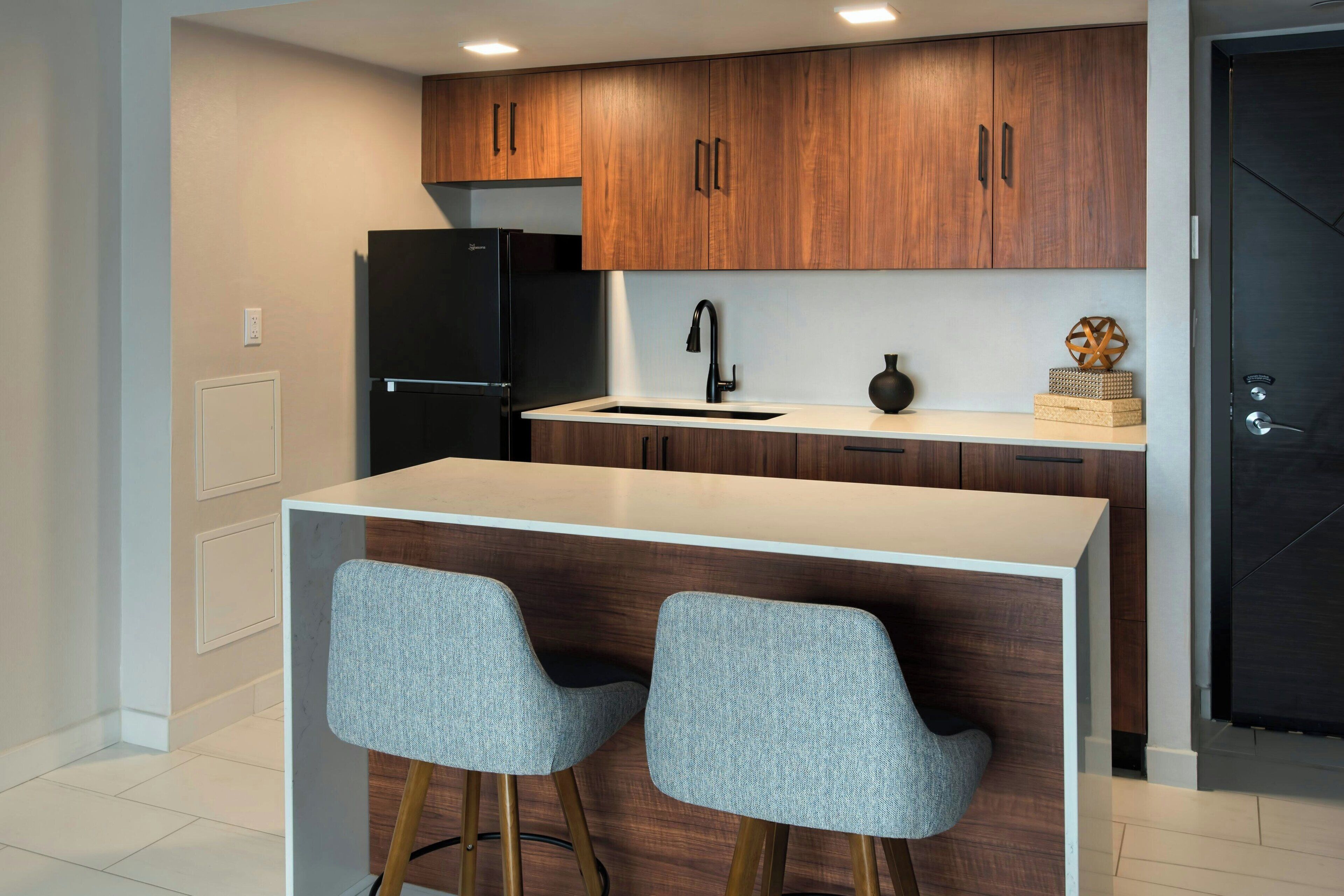 A modern kitchen with wooden cabinets, a black refrigerator, a white countertop, and two grey chairs.