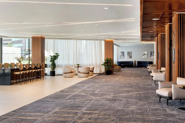 A modern hotel lobby with a long reception desk, several seating areas, and a large window with curtains.