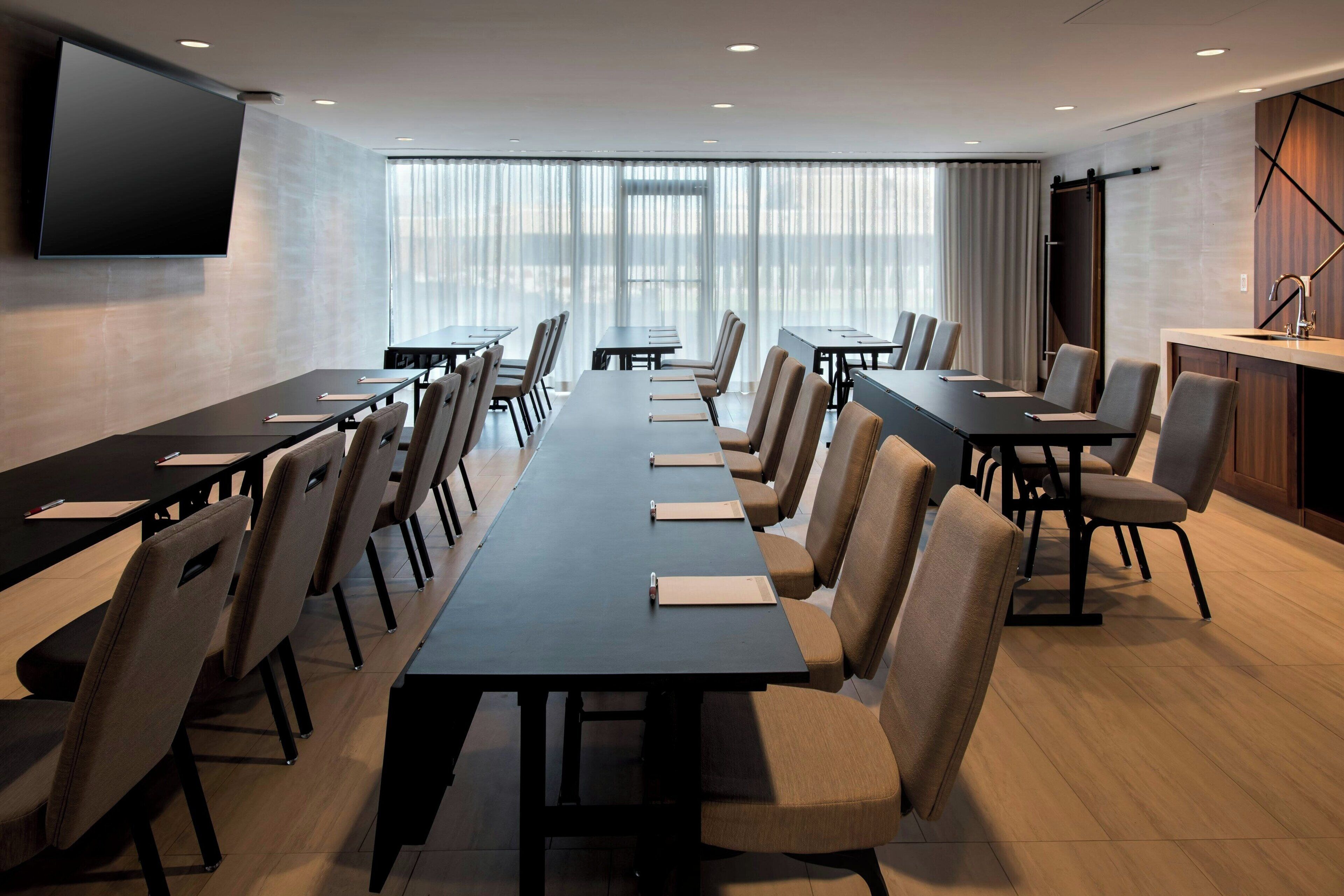 A conference room with long tables and chairs, a flat-screen TV mounted on the wall, and large windows with curtains.