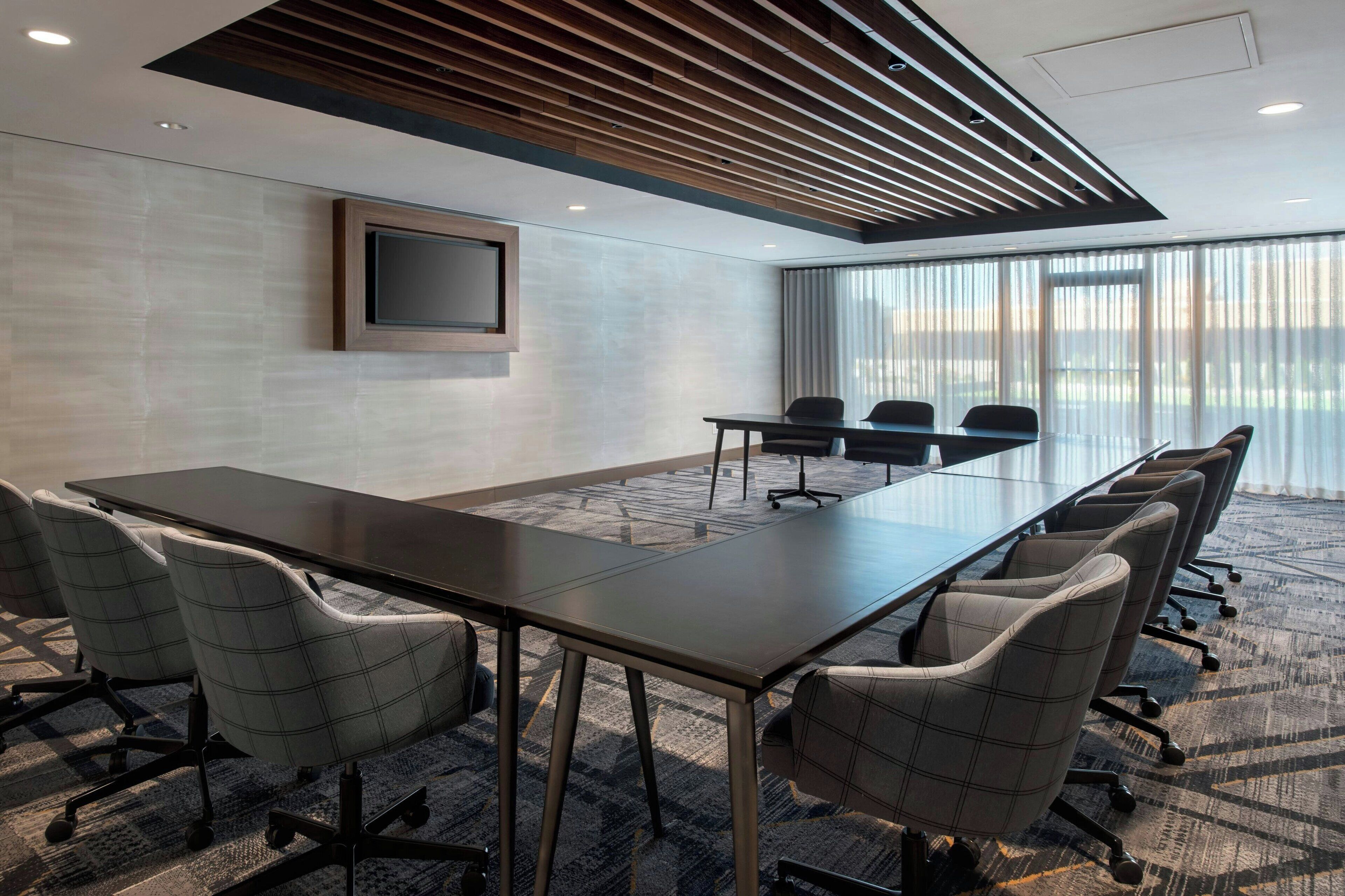 A conference room with a long table, six chairs, a flat-screen TV, and a large window with curtains.