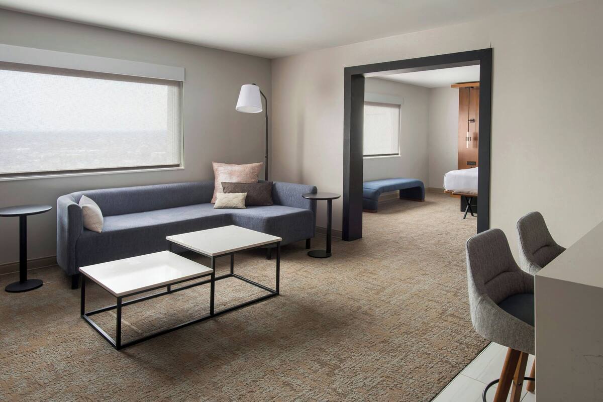 Una habitación de hotel moderna con un sofá gris, dos mesitas blancas, una mesita redonda pequeña y un sillón gris.