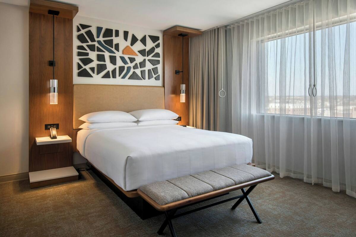 Una habitación de hotel moderna con una cama grande, un banco, una obra de arte en la pared y vistas a través de cortinas translúcidas.