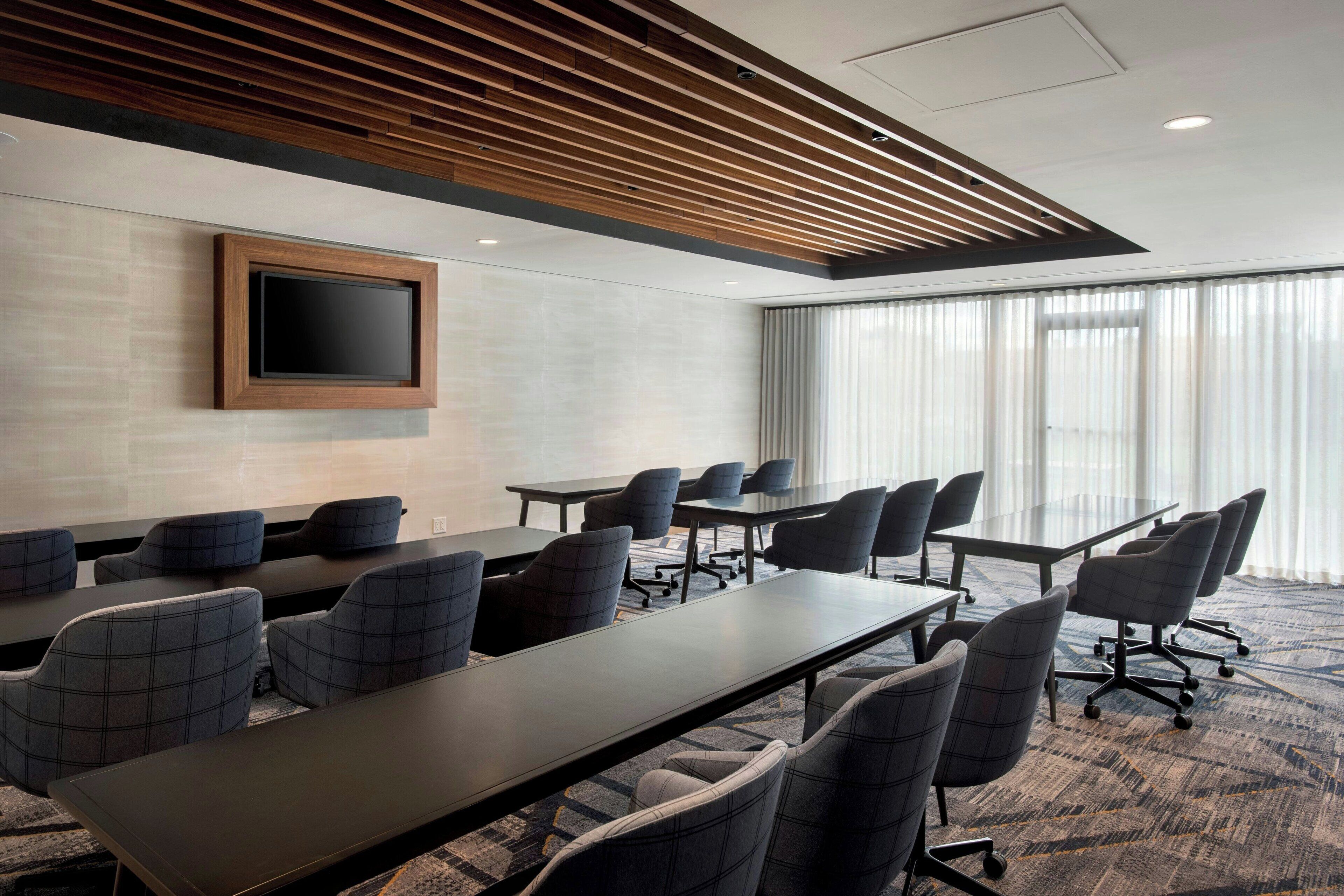 A conference room with a long table, multiple chairs, a flat-screen TV mounted on the wall, and large windows with sheer curtains.