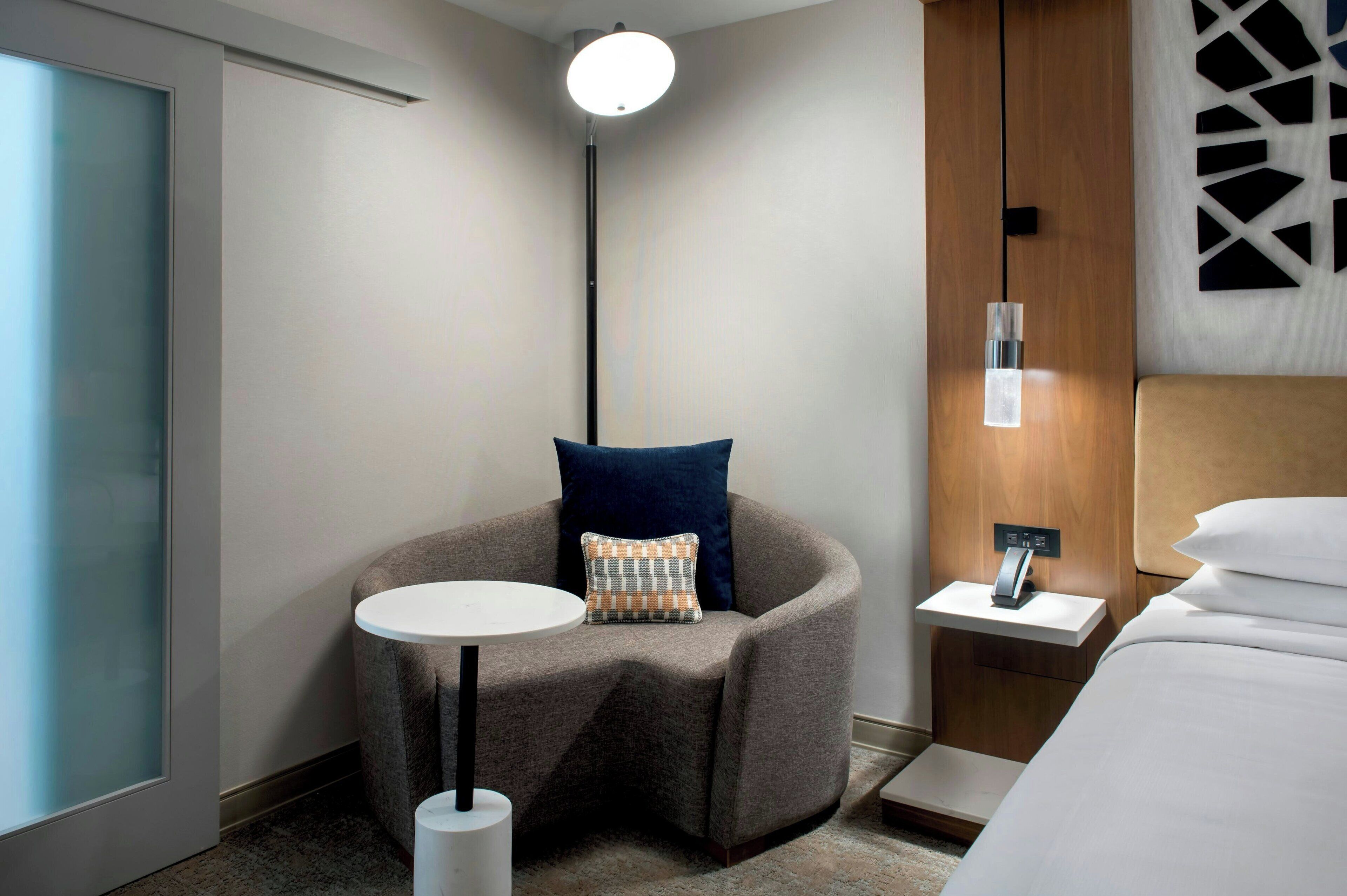 Una camera d'albergo moderna con una poltrona grigia a forma di ferro di cavallo, un piccolo tavolino, un comodino con una lampada, un letto con lenzuola bianche e un'ampia finestra con vetro satinato.