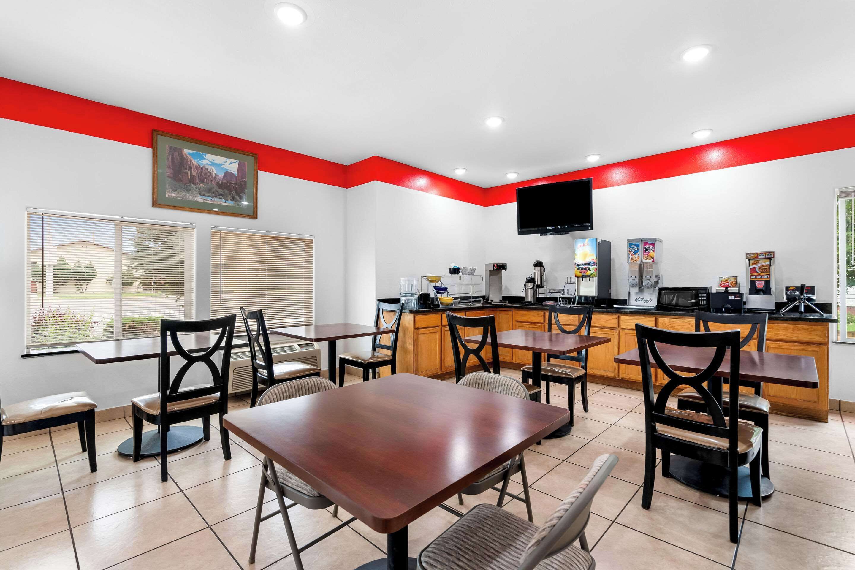 Un restaurante con una pared roja decorativa, mesas y sillas de madera, una zona de cocina y un televisor montado en la pared.