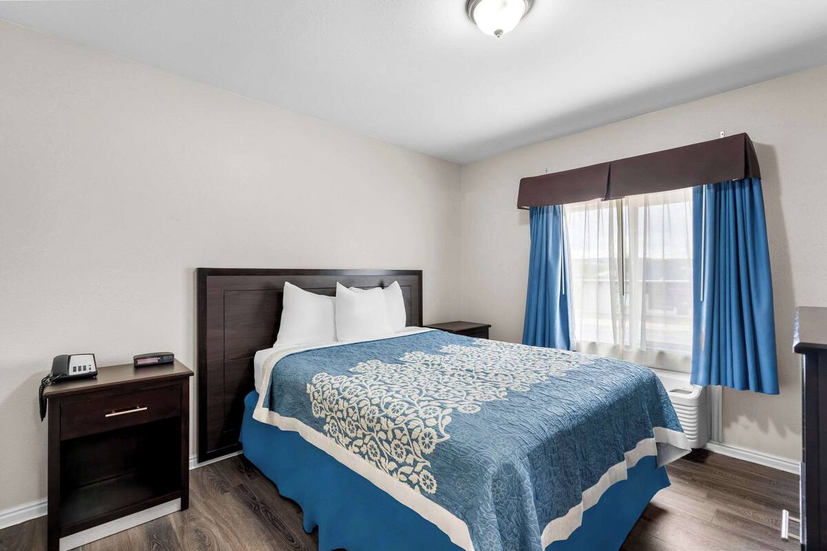 A hotel room with a bed, a bedside table with a phone, a window with blue curtains, and a wall-mounted heater.