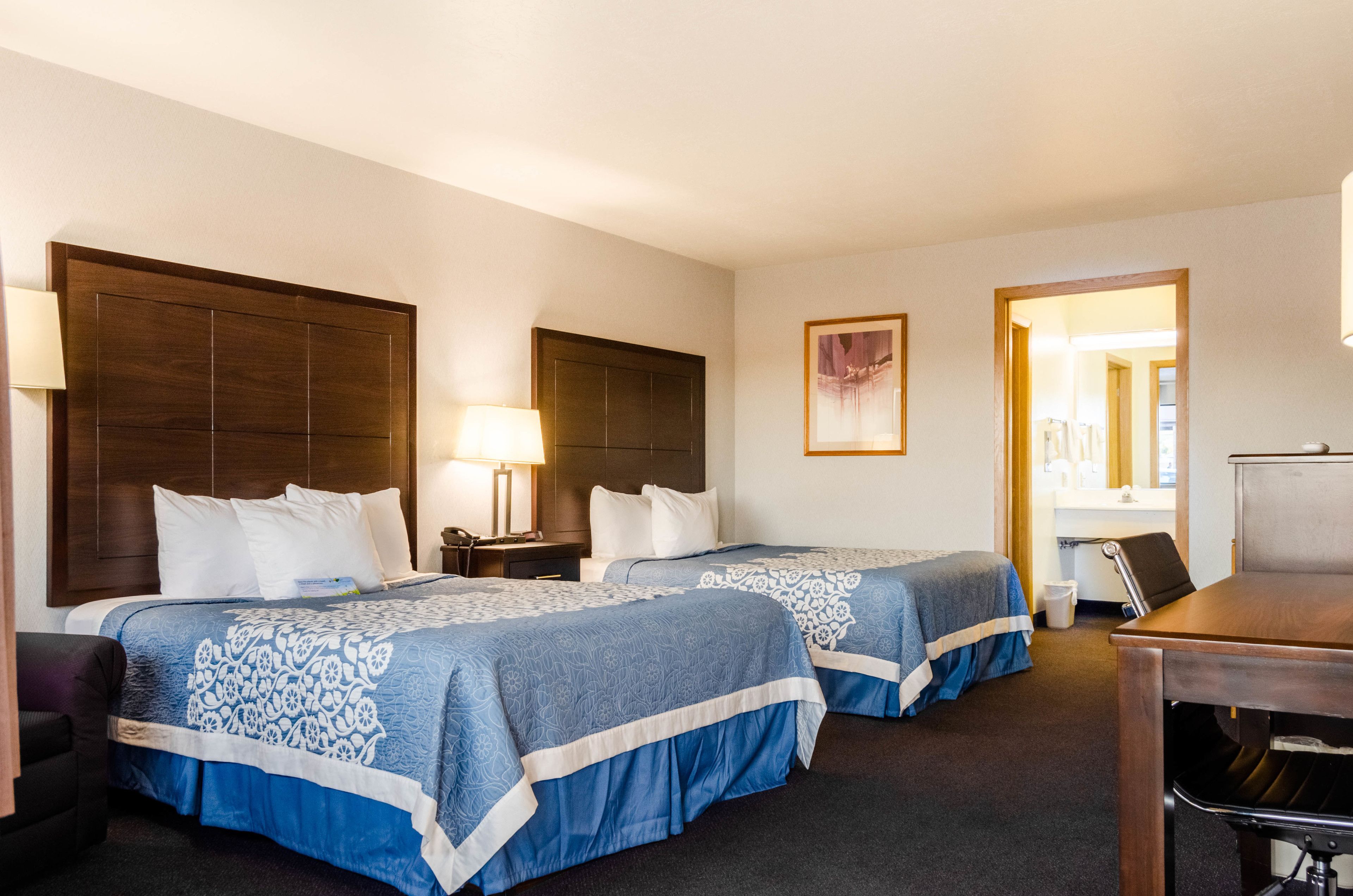 A hotel room with two beds, a desk, a chair, a lamp, and a picture on the wall.