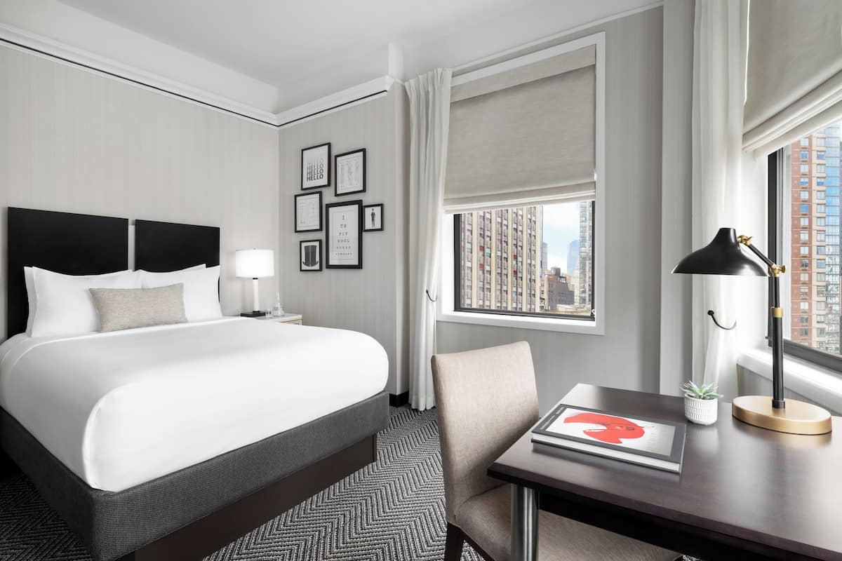 A hotel room with a large bed, a desk with a lamp, a chair, and framed pictures on the wall.