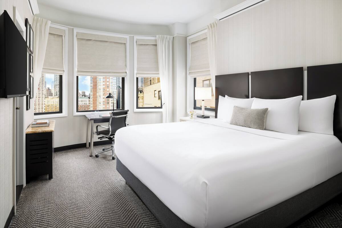A hotel room with a large bed, a desk, and a chair. There are windows with curtains, a flat-screen TV mounted on the wall, and a carpeted floor.