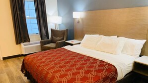 Una habitación de hotel con cama, silla, mesita de noche, lámpara y equipo de aire acondicionado.