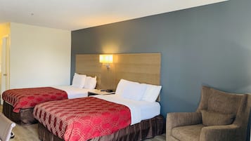 A hotel room with two beds, a brown armchair, and a nightstand with a lamp.