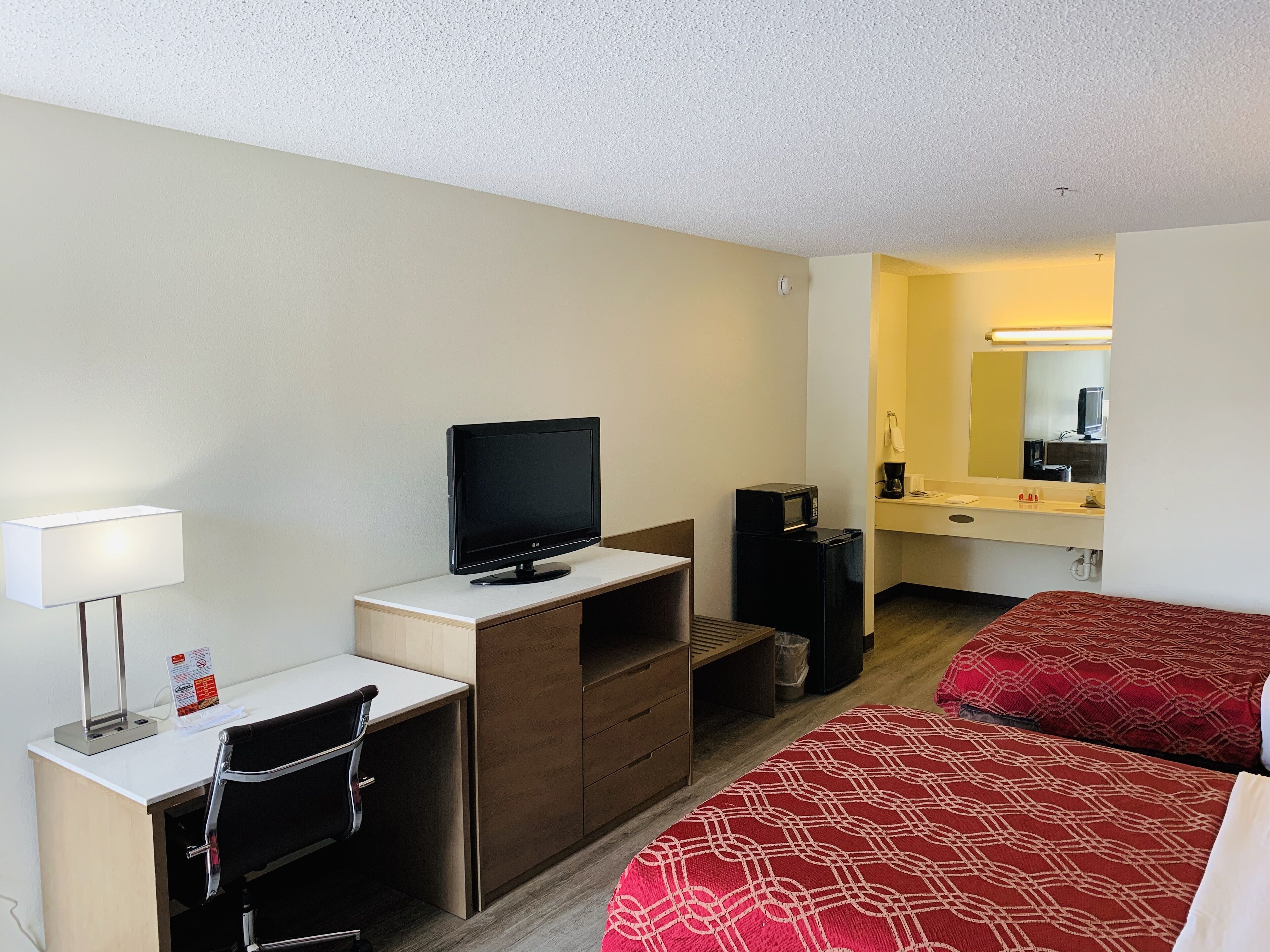A hotel room with a desk, a television, a microwave, a bed with patterned bedding, and a bathroom with a mirror and sink.