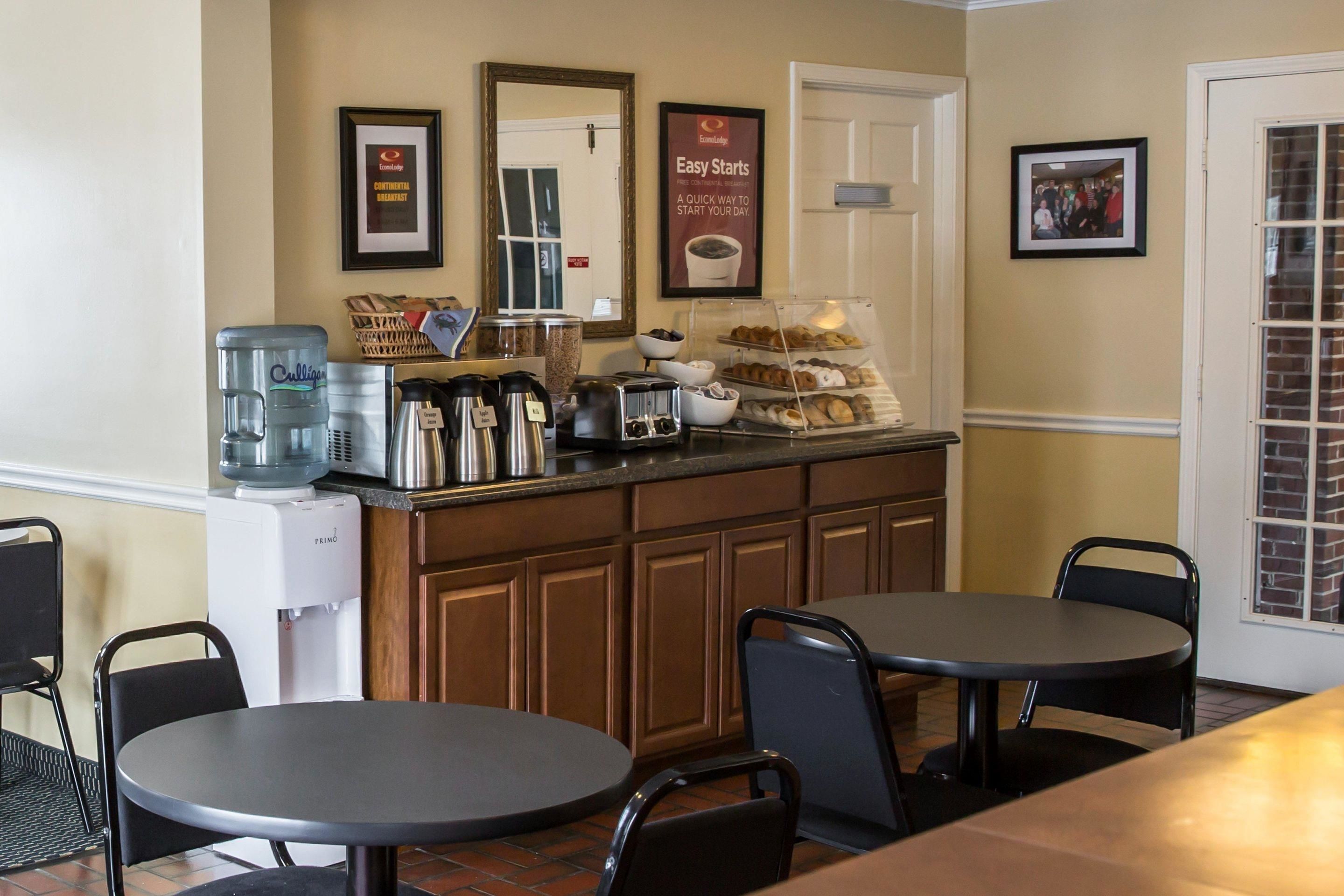 A breakfast bar with coffee makers, a water dispenser, and a display of pastries. There are two tables and chairs in the foreground.