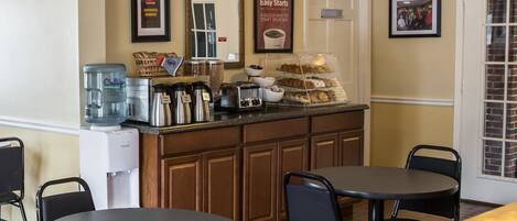 A breakfast bar with coffee makers, a water dispenser, and a display of pastries. There are two tables and chairs in the foreground.