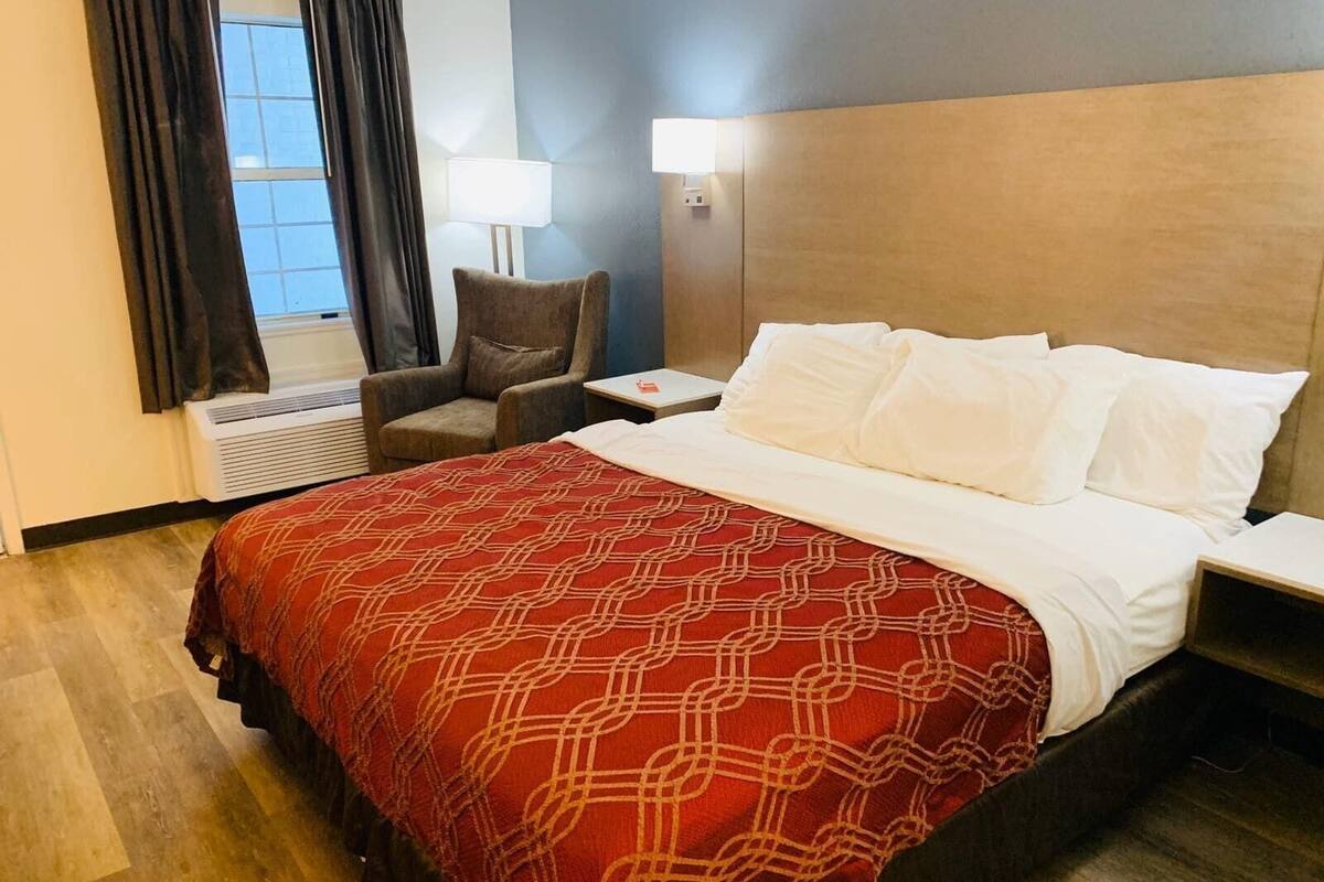 A hotel room with a bed, a chair, a nightstand, a lamp, and a window with curtains.