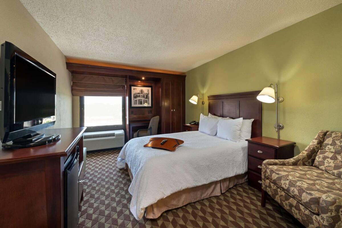 A hotel room with a bed, a desk with a TV, a chair, and a lamp.