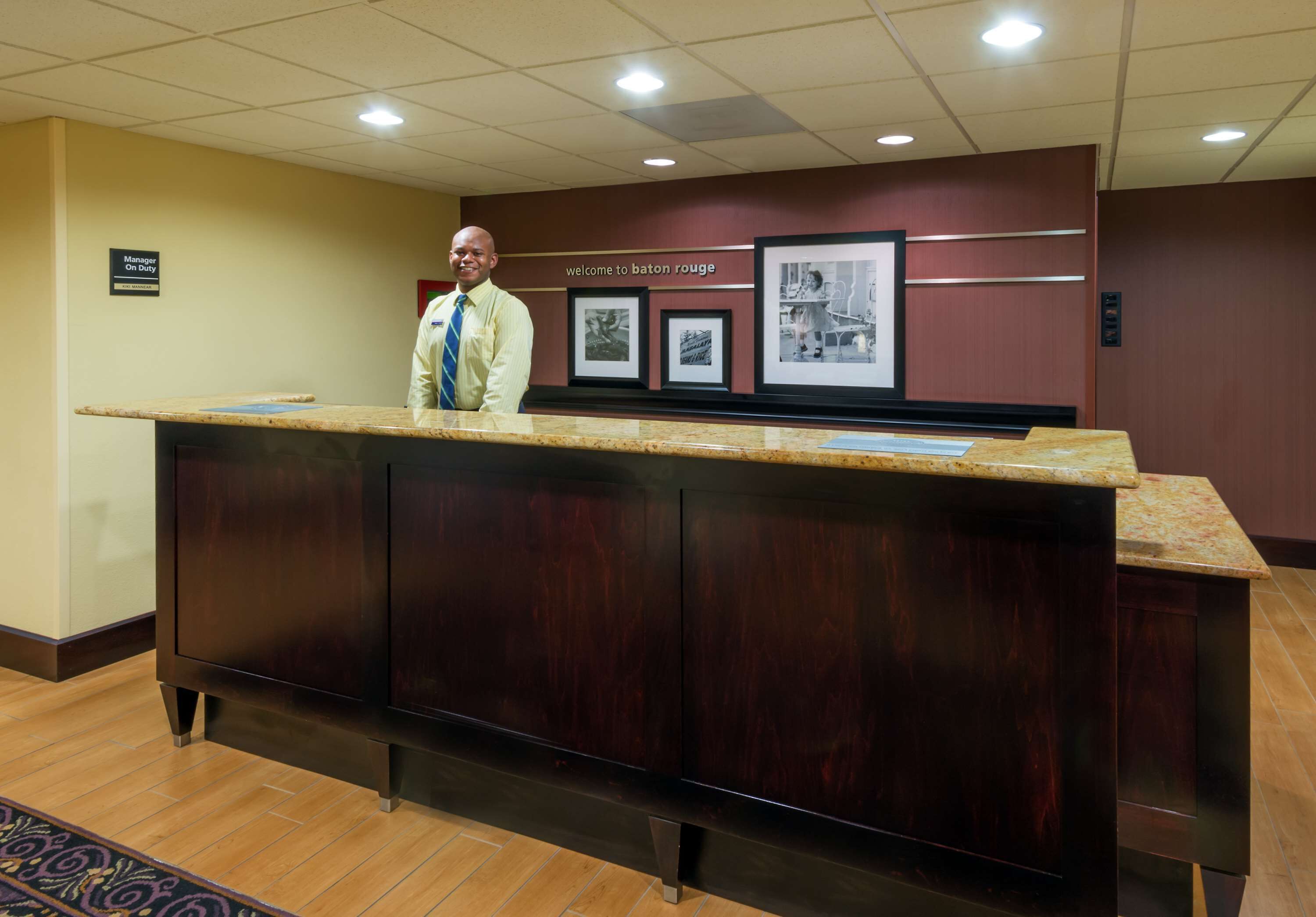 Een receptiebalie met een man erachter, ingelijste foto's aan de muur en een bord met de tekst 'Welcome to Baton Rouge'.