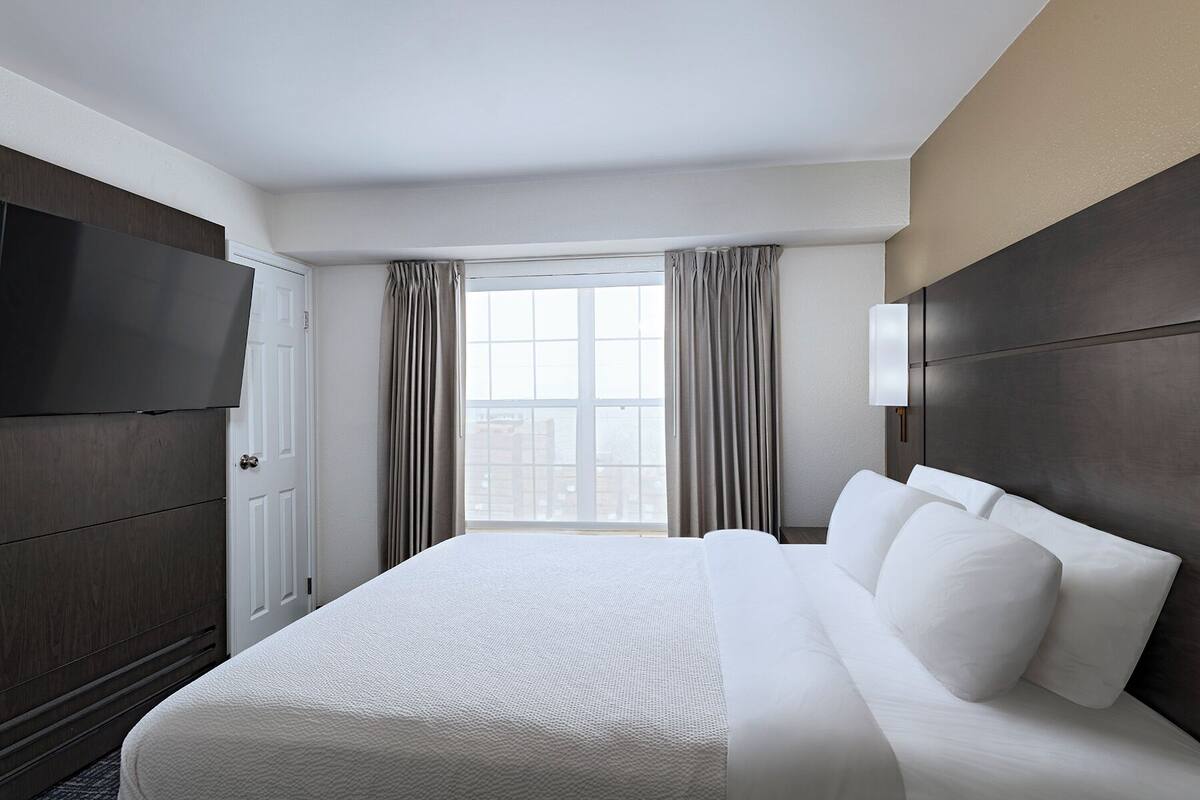 Een hotelkamer met een groot bed, een televisie, een raam met gordijnen en een donker hoofdeinde.