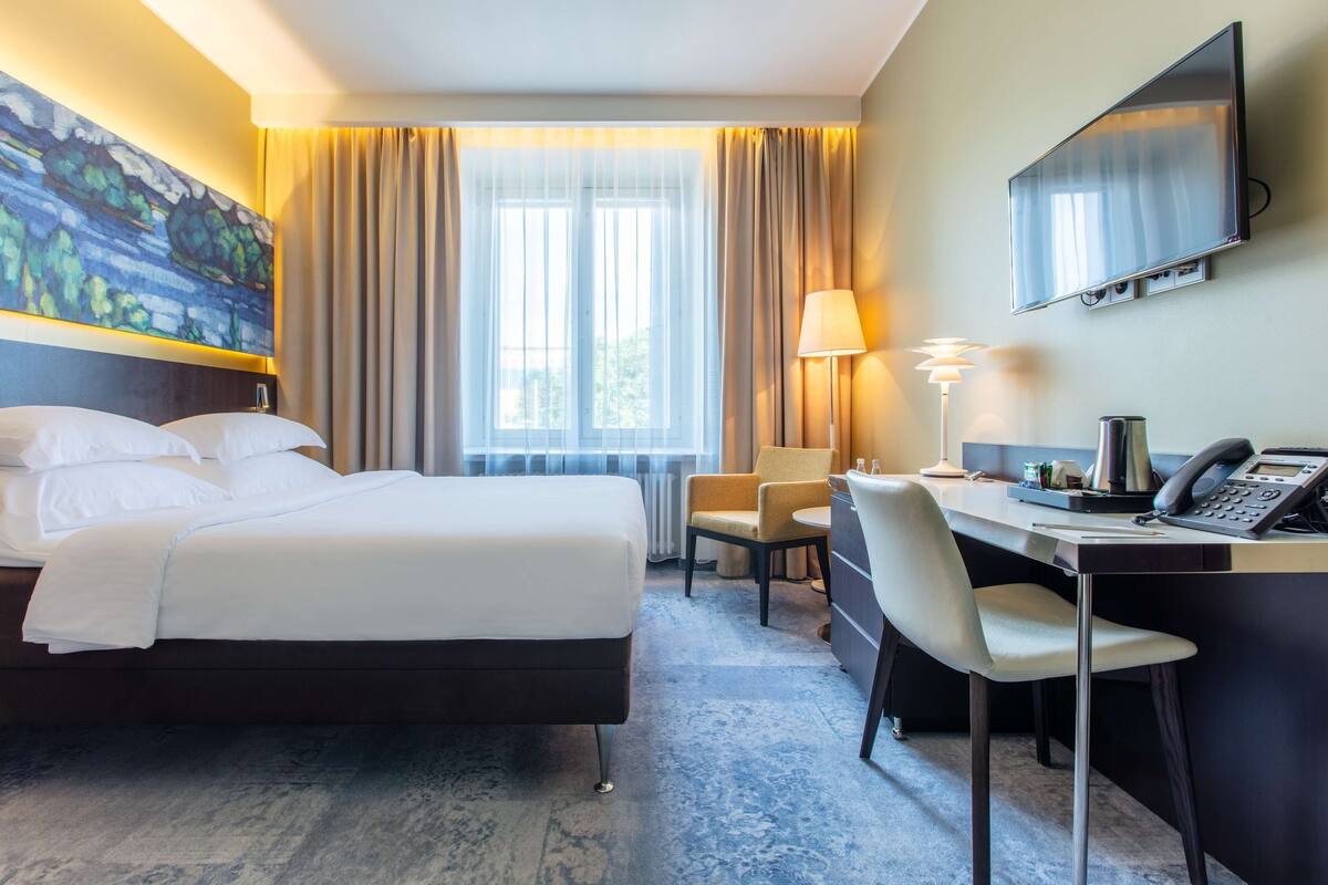 A hotel room with a bed, a desk with a telephone, a chair, a lamp, and a painting on the wall.