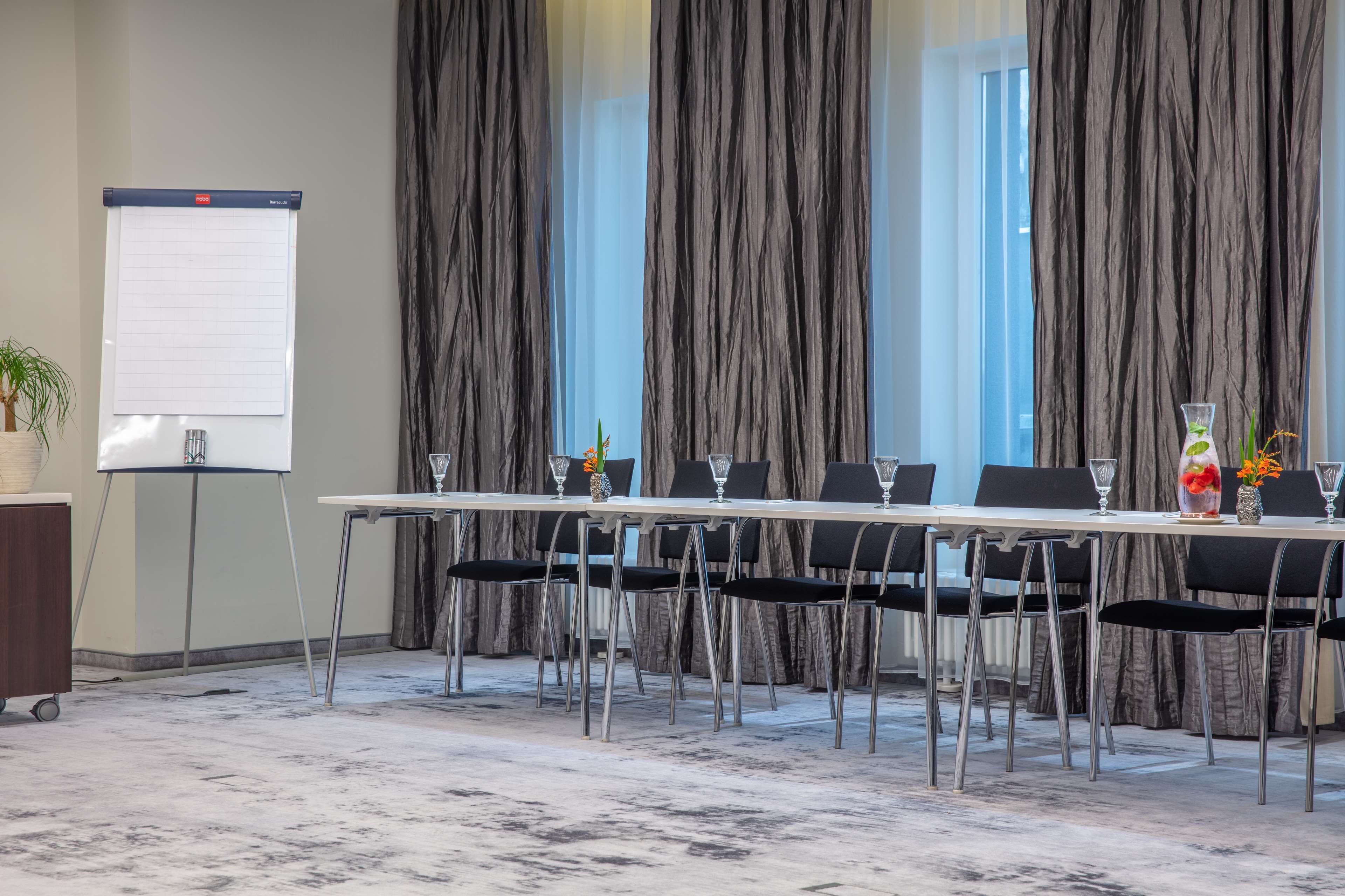 Ein Konferenzraum mit einem langen Tisch, schwarzen Stühlen, einer Whiteboard-Wand und Blick durch große Vorhänge.