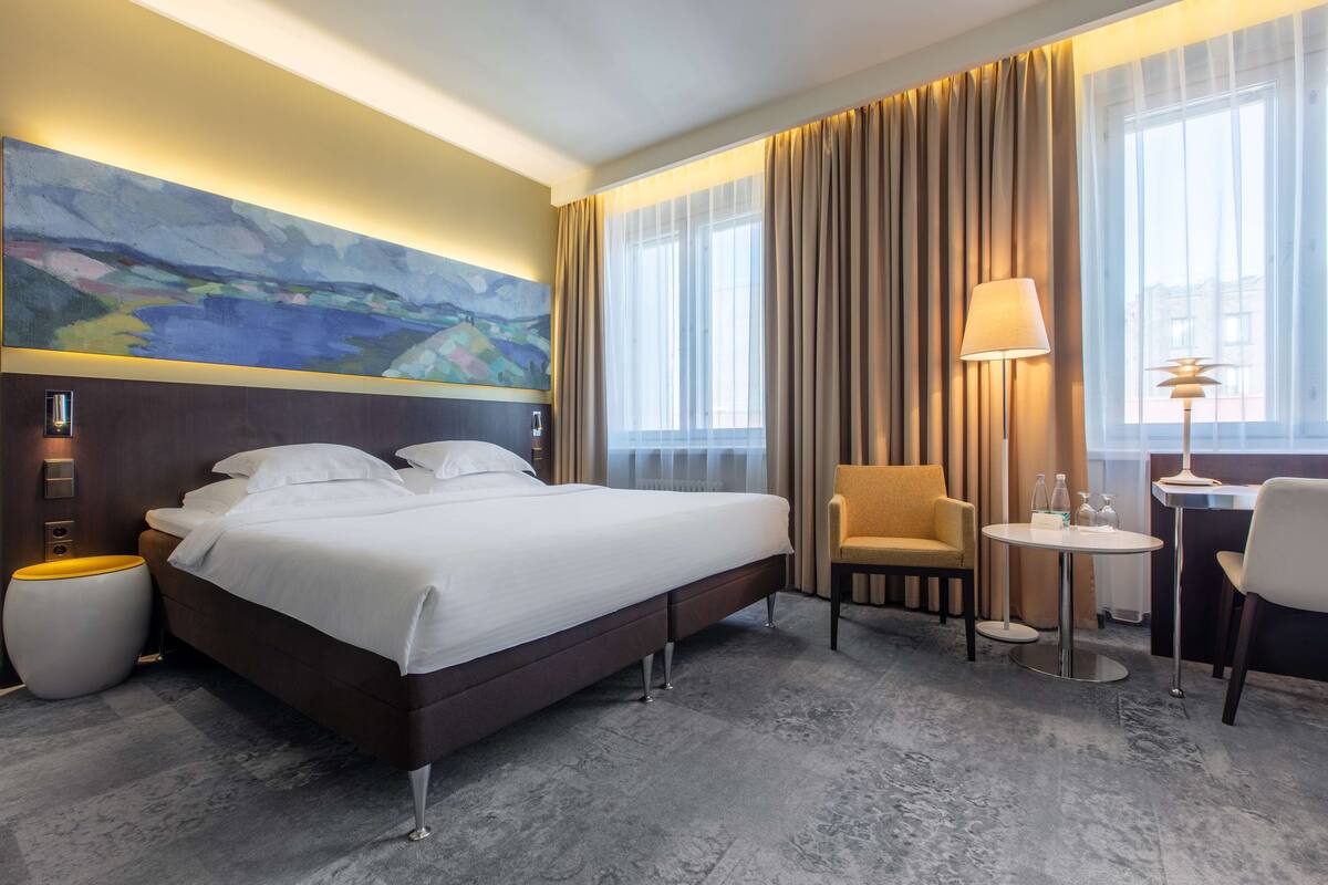 A hotel room with a large bed, a chair, a small table, and a painting on the wall.