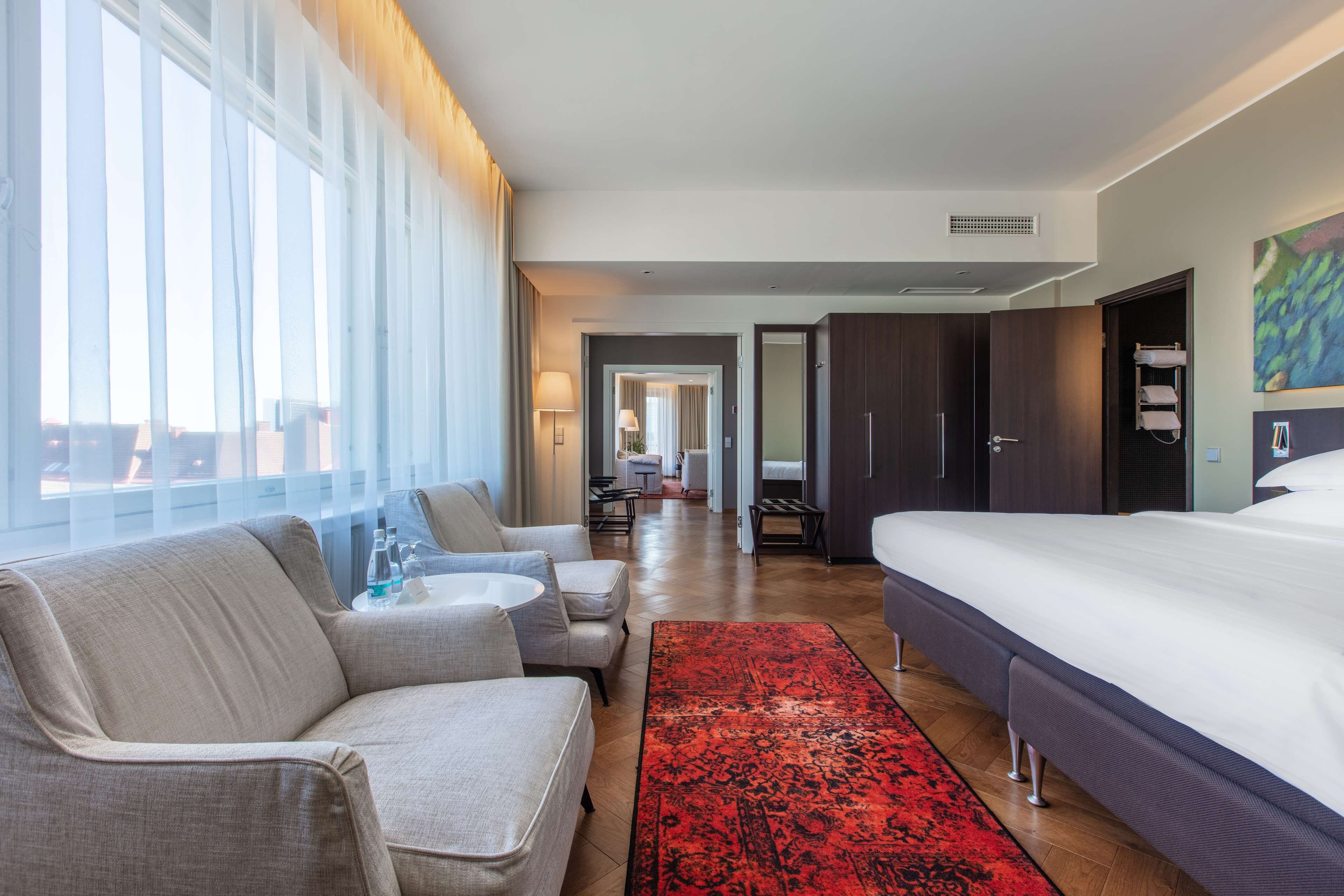 A hotel room with a large bed, two armchairs, a red rug, and a view of the city through large windows.
