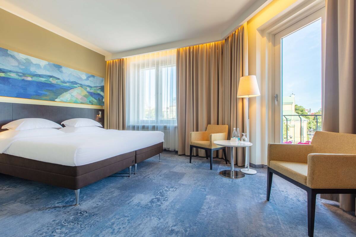 A hotel room with a large bed, a chair, a small table, and a painting on the wall.