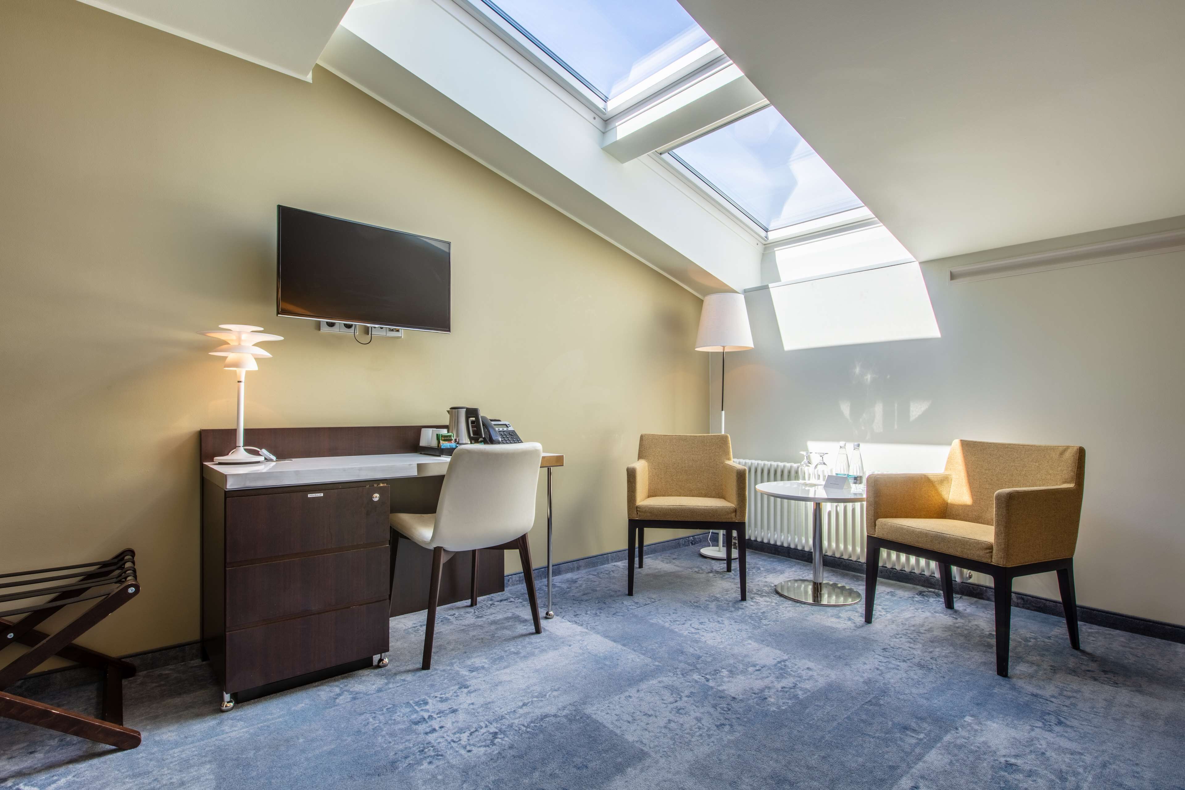 A hotel room with a skylight, a desk with a chair, a sofa, a lamp, and a TV mounted on the wall.