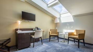 A hotel room with a skylight, a desk with a chair, a sofa, a lamp, and a TV mounted on the wall.