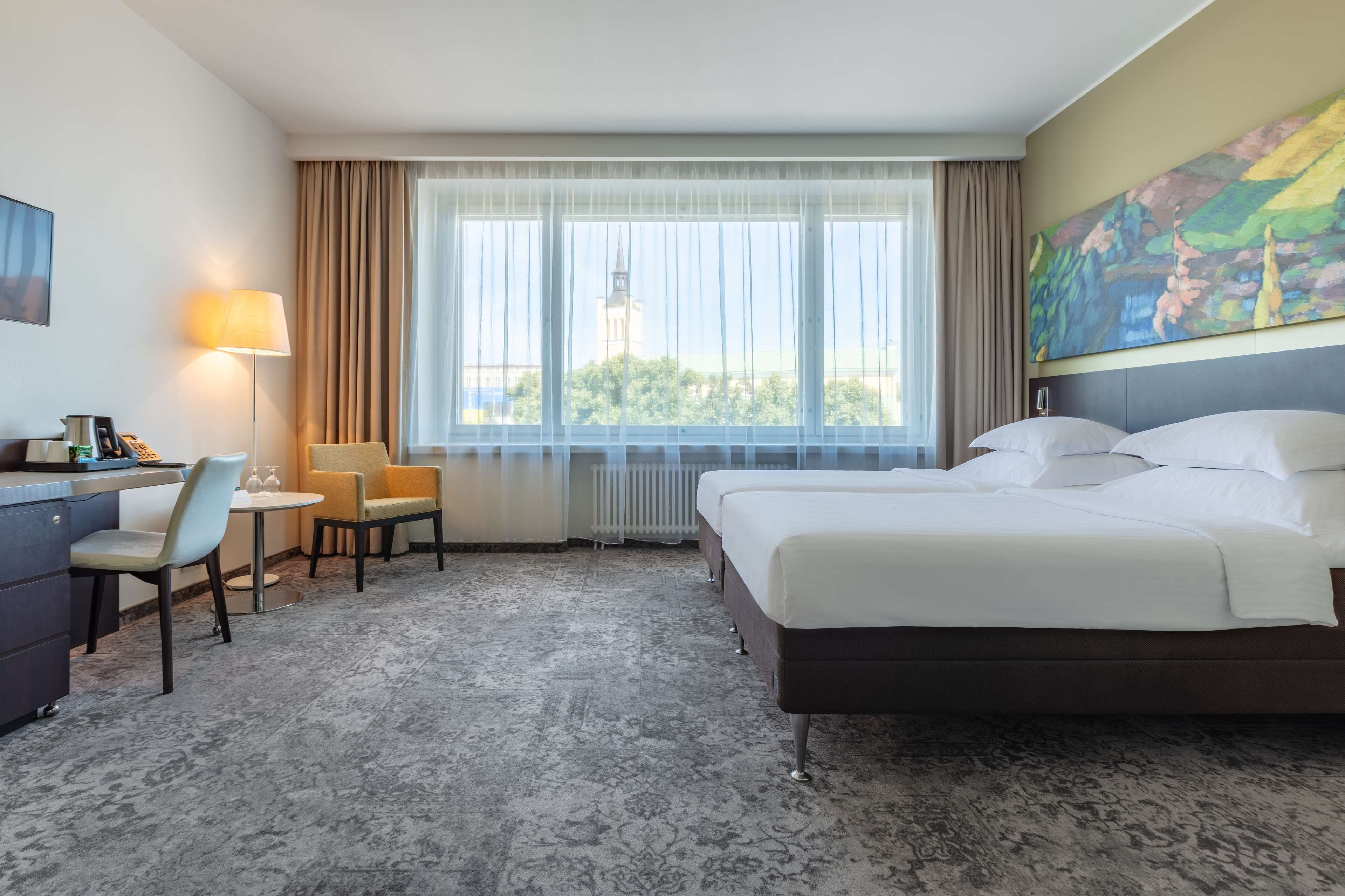 Habitación de hotel con cama, dos sillas, un escritorio, una lámpara y un ventanal con vistas a un edificio.