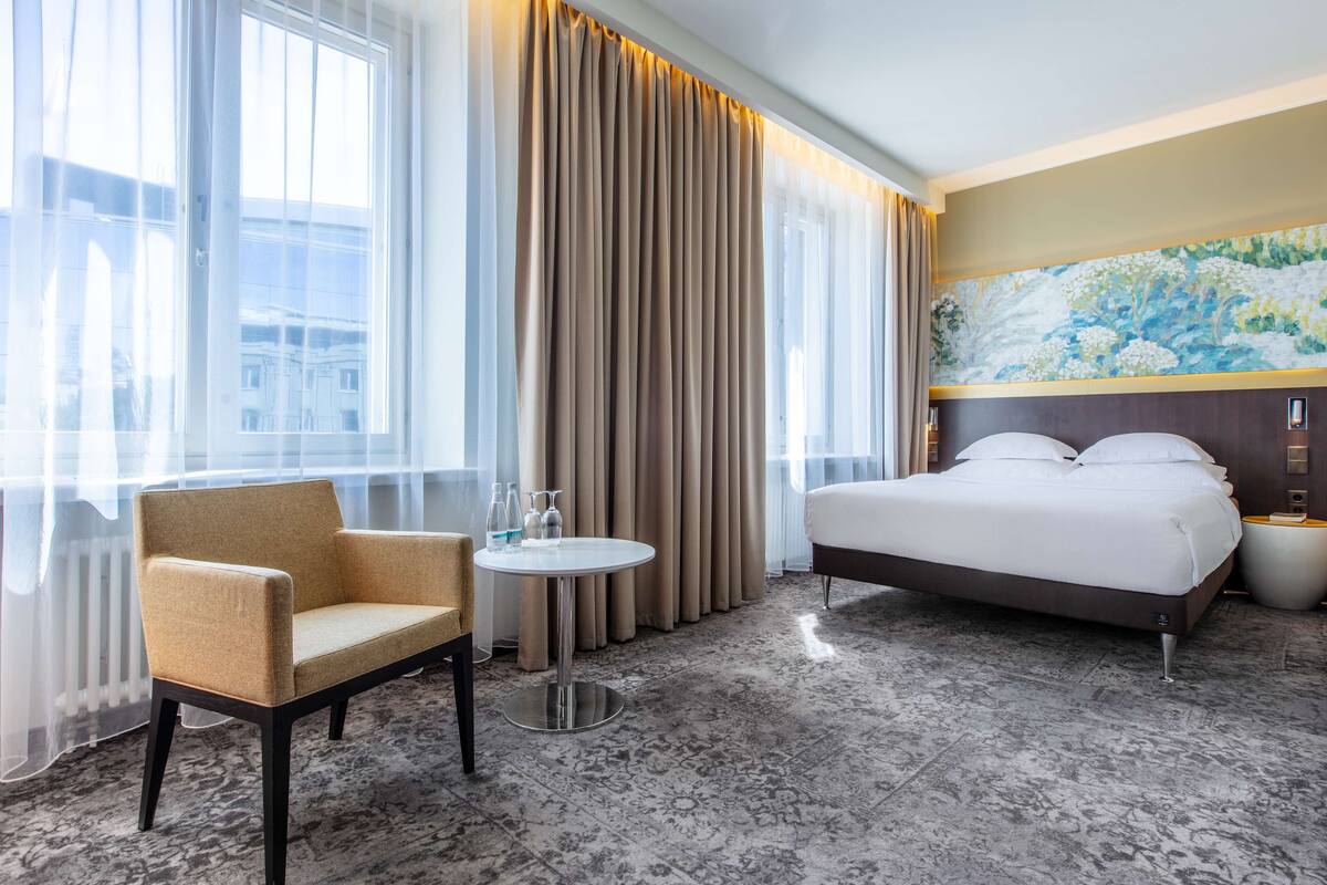 A hotel room with a bed, a chair, a round table, and a large window with curtains.