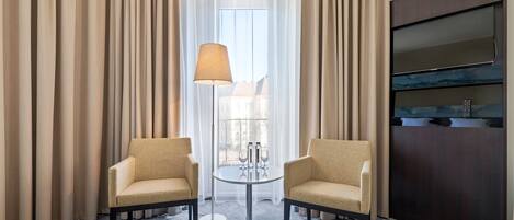 A hotel room with two beige armchairs, a small round table with a lamp, and a large dark wood cabinet.