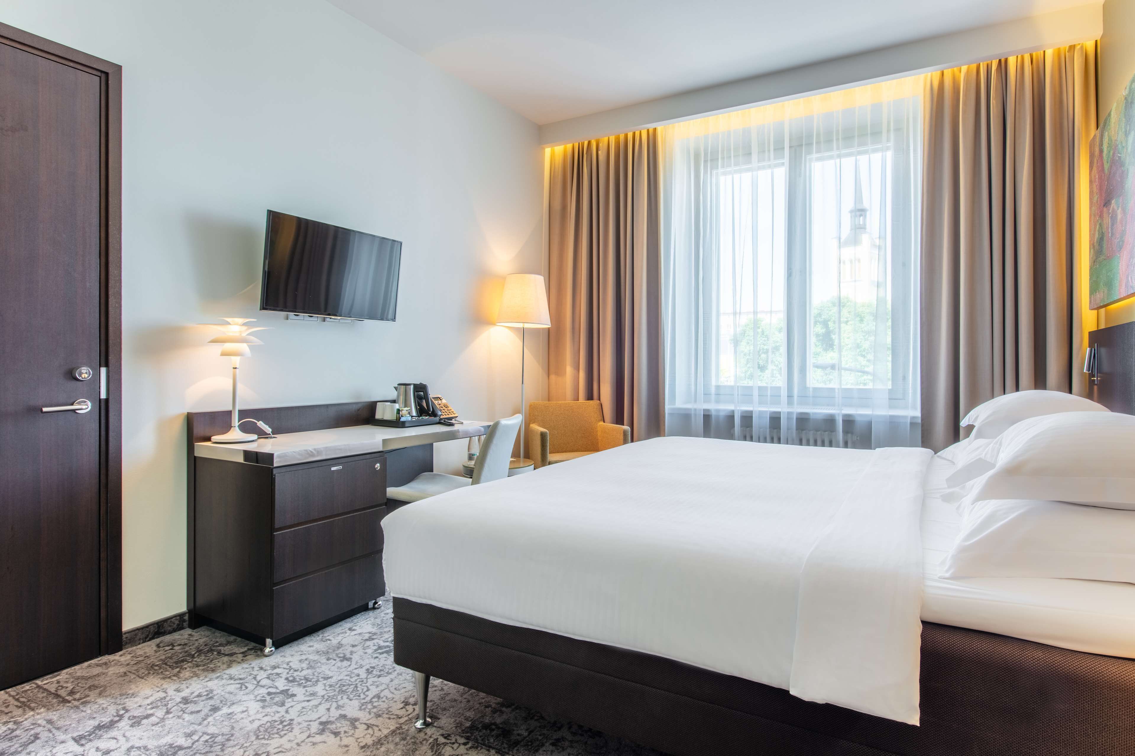 Een hotelkamer met een bed, een bureau met lamp, een stoel, een televisie aan de muur en een raam met gordijnen.