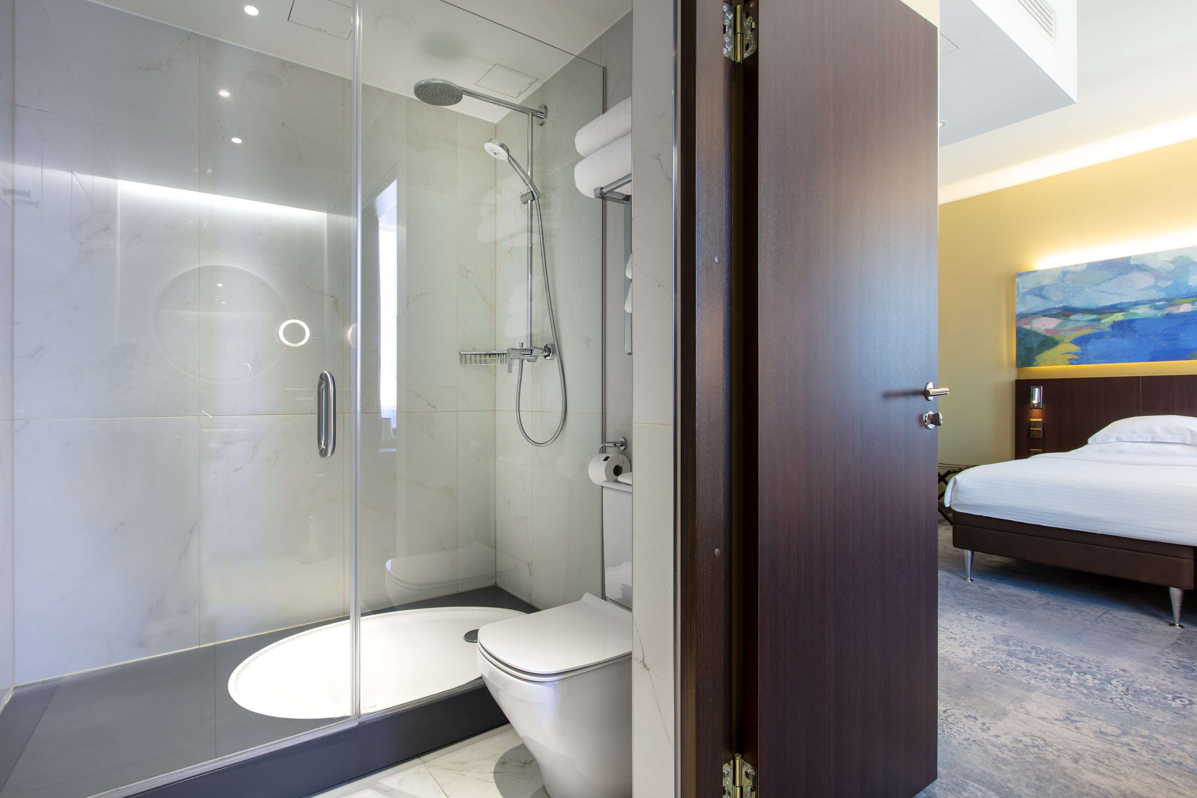 A modern bathroom with a glass-enclosed shower, a toilet, and a bed with a wooden headboard.