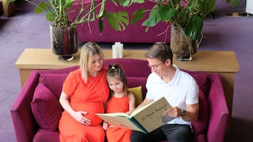 A family on a purple sofa with a book titled 'Le Petit Prince' and potted plants in the background.