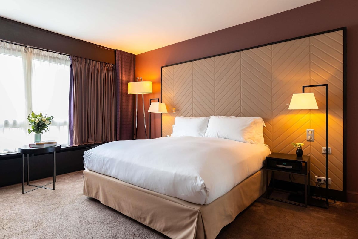 Habitación de hotel con una cama grande, dos mesitas de noche, una lámpara, un florero con flores y vistas a la ciudad a través de una ventana.