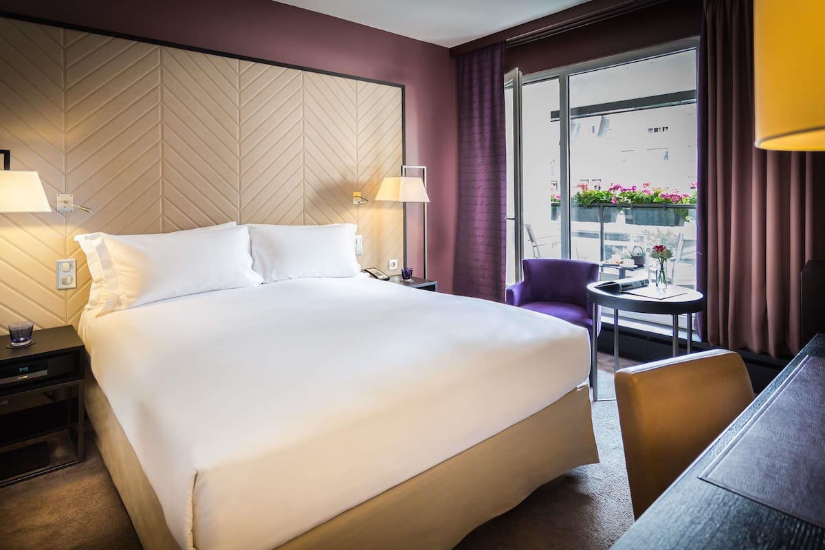 Habitación de hotel con una cama grande, dos lámparas de noche, una mesita de noche, una pequeña mesa con una silla y vistas al exterior por una ventana.